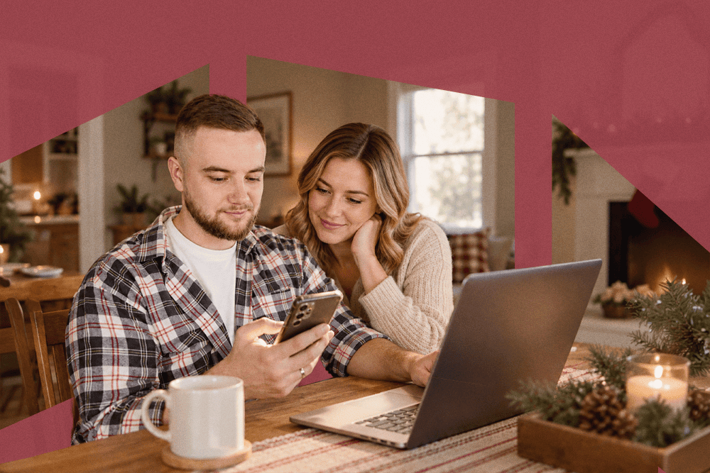 Couple sitting at a dining table with a laptop, looking at a smartphone together in a cozy home