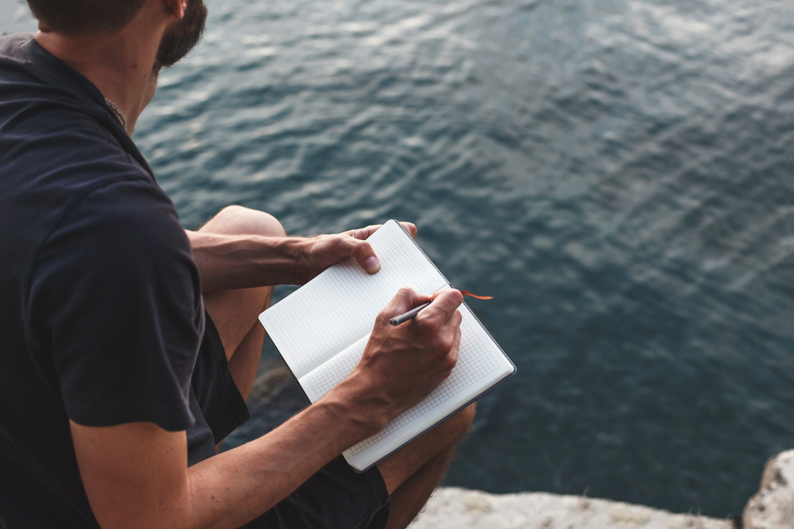 man sits on a rocky shore by water, writing in notebook. he is immersed in his thoughts, journaling peacefully in nature. calm waves, quiet atmosphere. Concept: mindfulness, solitude, mental health.
