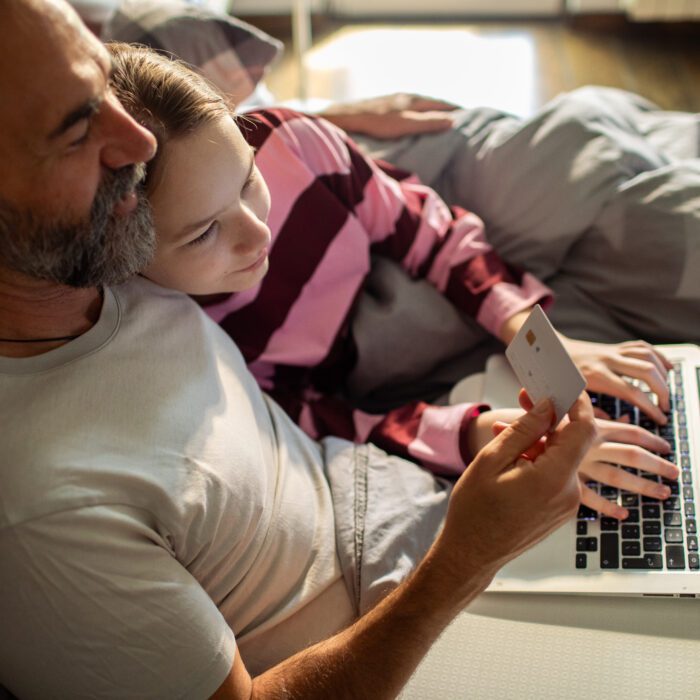 A man and child sit together on a bed, sharing a laptop. The child types while the man holds a credit card, suggesting online shopping. Cozy and focused.