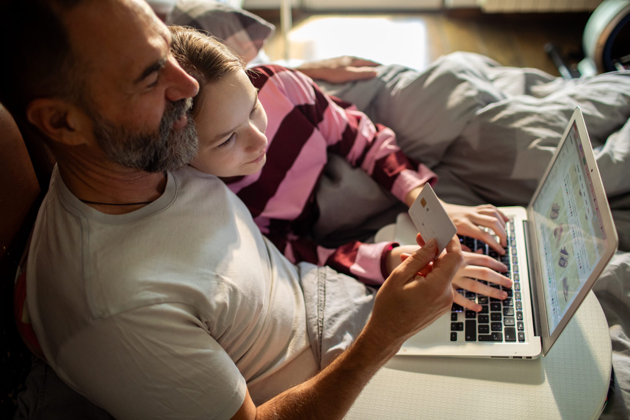 A man and child sit together on a bed, sharing a laptop. The child types while the man holds a credit card, suggesting online shopping. Cozy and focused.