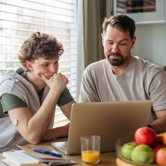 Father helping teenage son with diabetes with homework on laptop in kitchen.