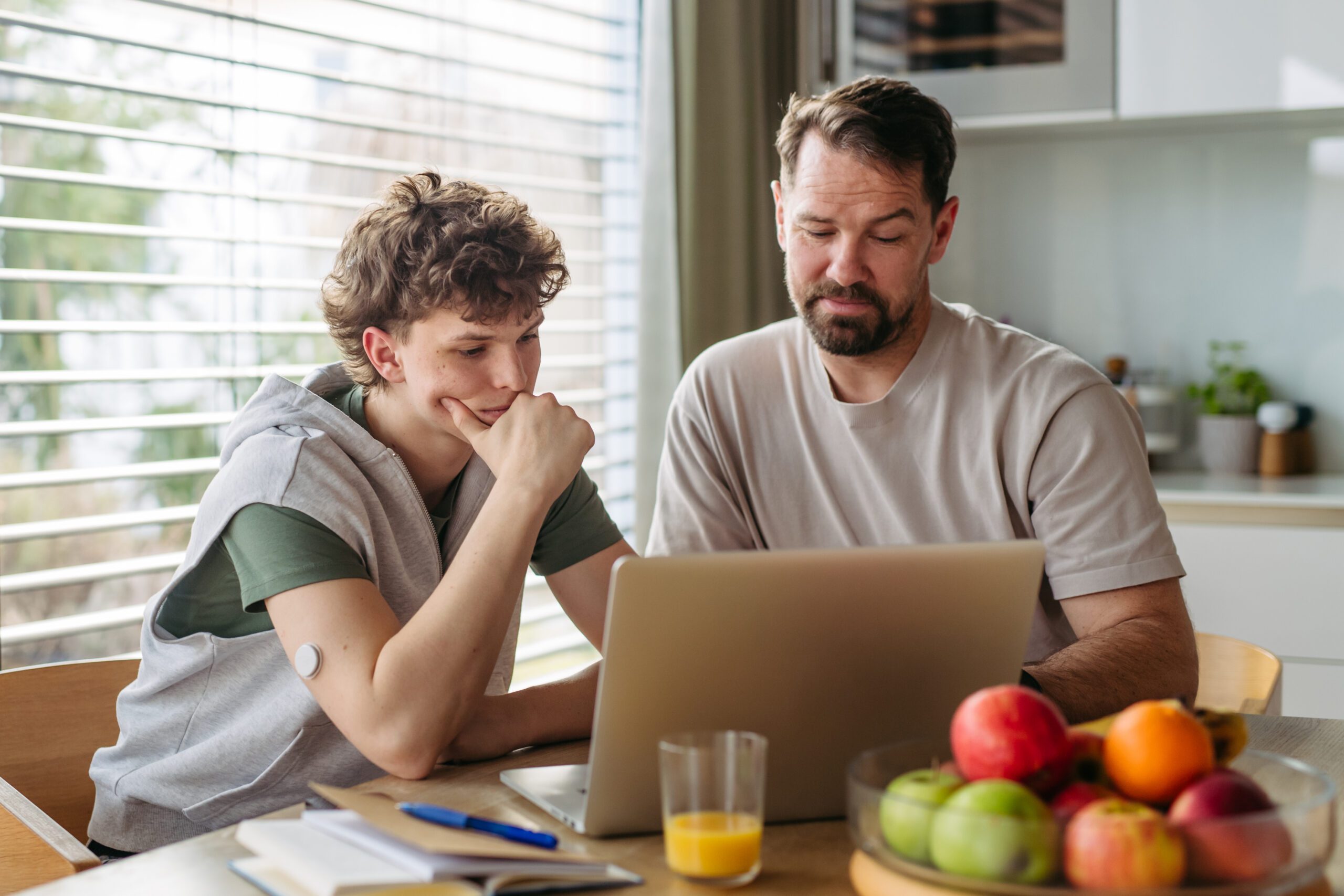 Father helping teenage son with diabetes with homework on laptop in kitchen.