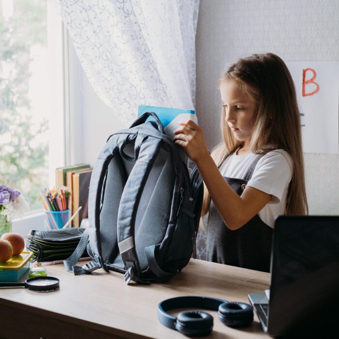 A young girl packs a blue backpack at a desk near a window with flowers and art supplies. A laptop and headphones sit nearby, conveying a studious vibe.