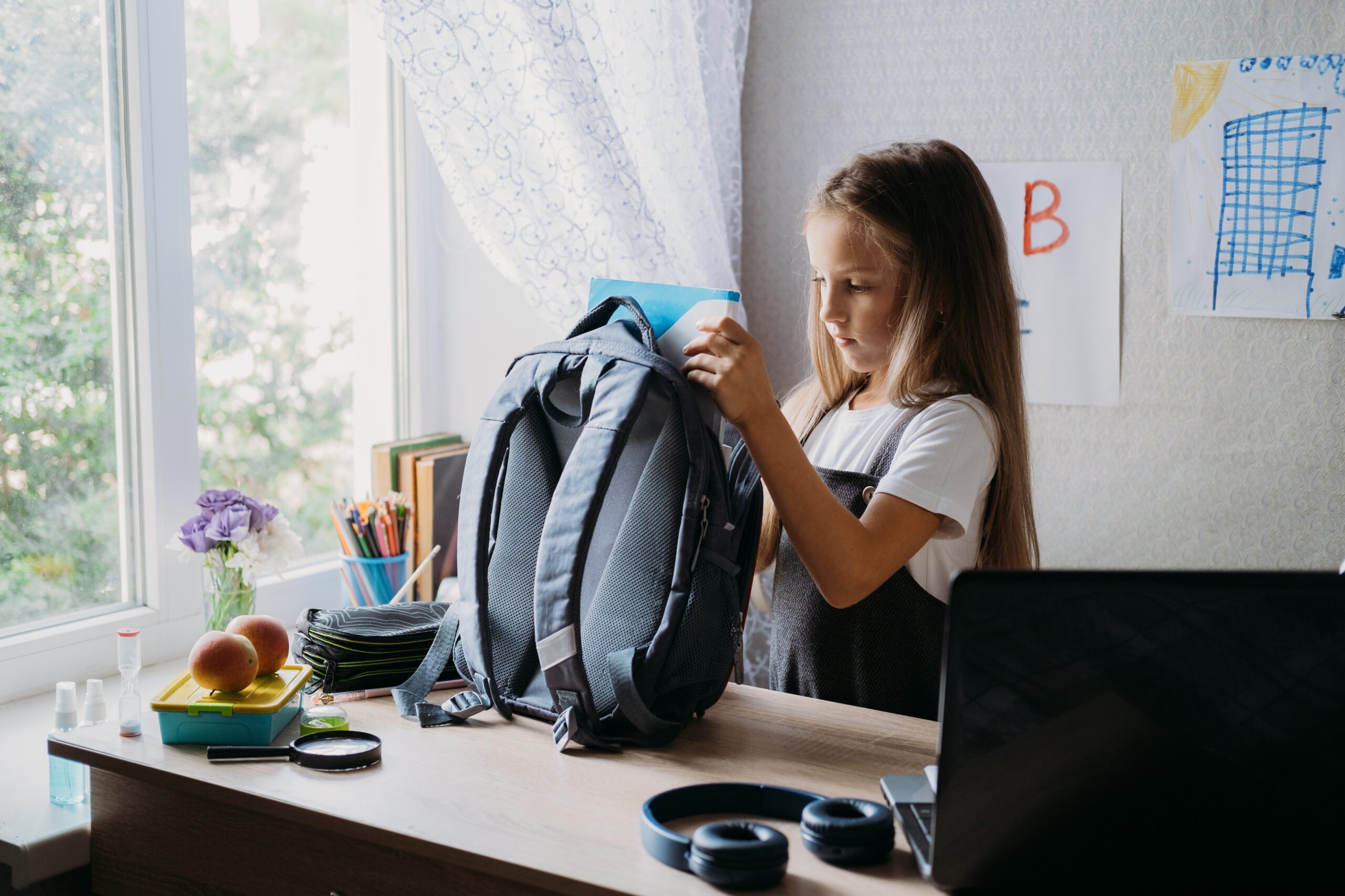 A young girl packs a blue backpack at a desk near a window with flowers and art supplies. A laptop and headphones sit nearby, conveying a studious vibe.