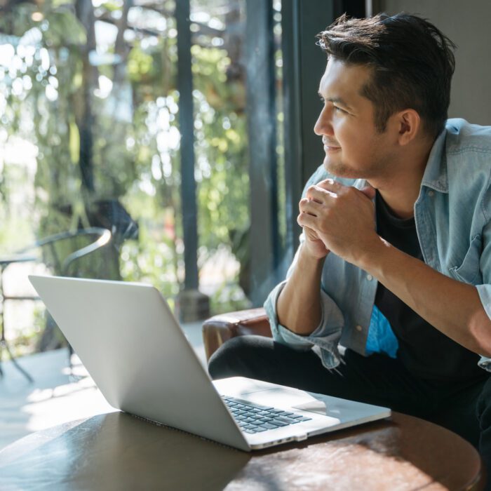Man in a denim shirt sits thoughtfully by a laptop, gazing out a window. Sunlight and greenery create a serene, reflective atmosphere in a modern setting.