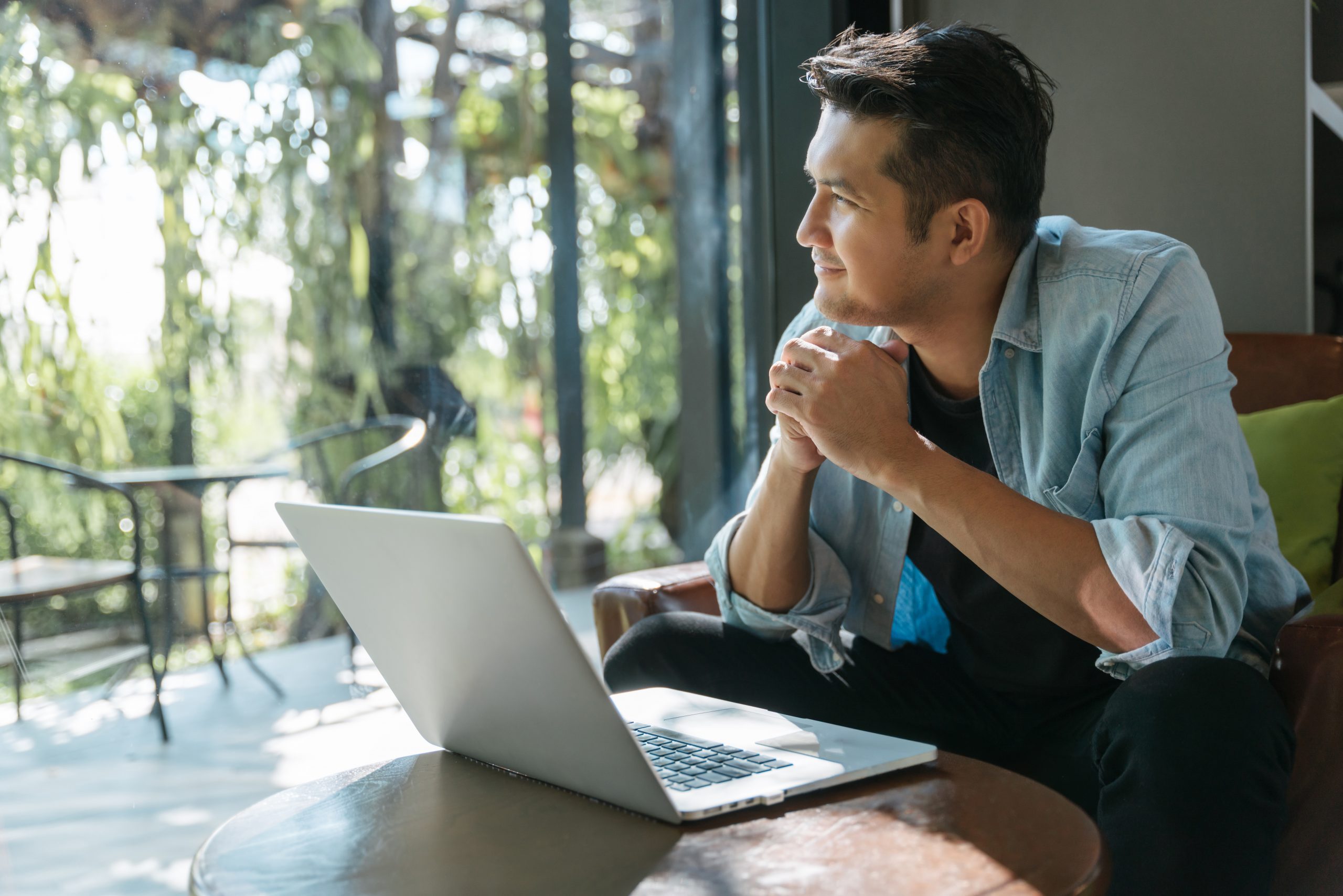 Man in a denim shirt sits thoughtfully by a laptop, gazing out a window. Sunlight and greenery create a serene, reflective atmosphere in a modern setting.