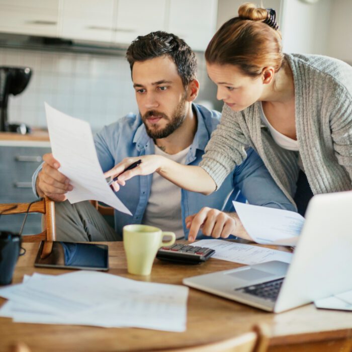 A couple reviews paperwork at a kitchen table, looking focused. Numerous documents, a laptop, calculator, and coffee cup indicate they are budgeting finances.